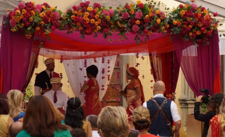 Crimson and magenta draped maṇḍapa crowned with pink and orange roses against an ochre villa wall, the wedding party within and guests seated close in front