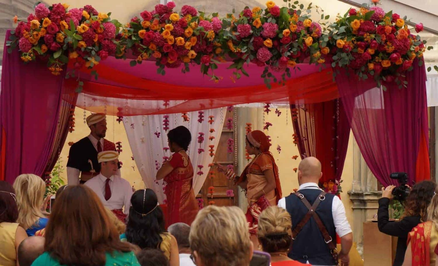 Crimson and magenta draped maṇḍapa crowned with pink and orange roses against an ochre villa wall, the wedding party within and guests seated close in front