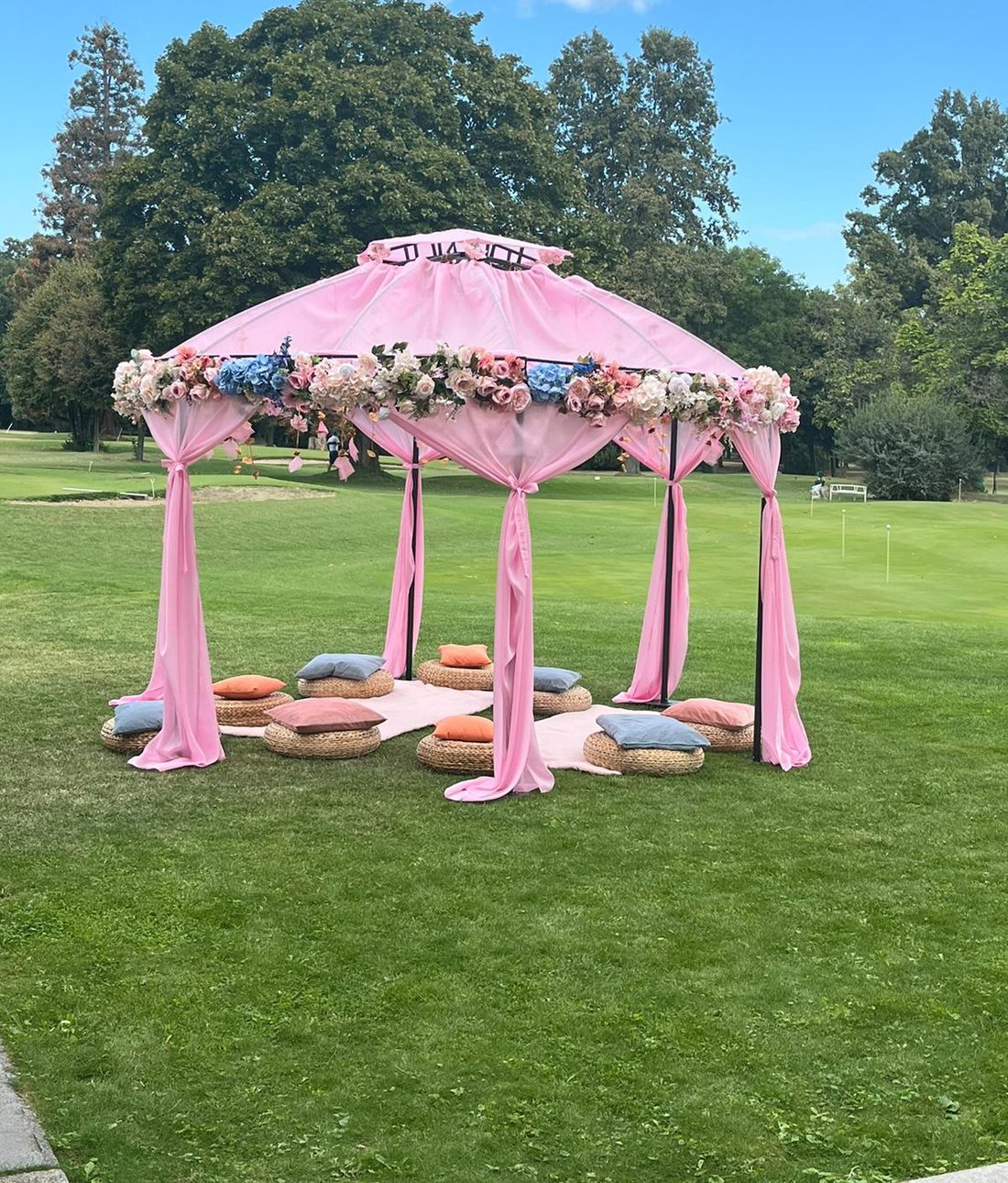 Pink draped maṇḍapa with rose and hydrangea garlands standing on a European lawn, floor cushions arranged beneath