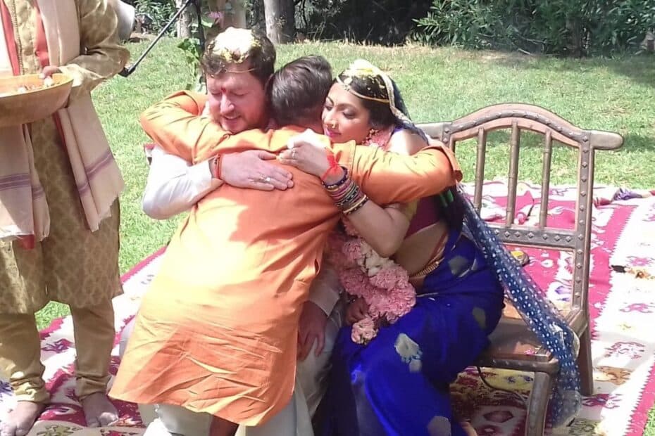 Bride in blue and groom in saffron embracing an elder on the ceremonial mat, with a plate of akṣata held ready beside them