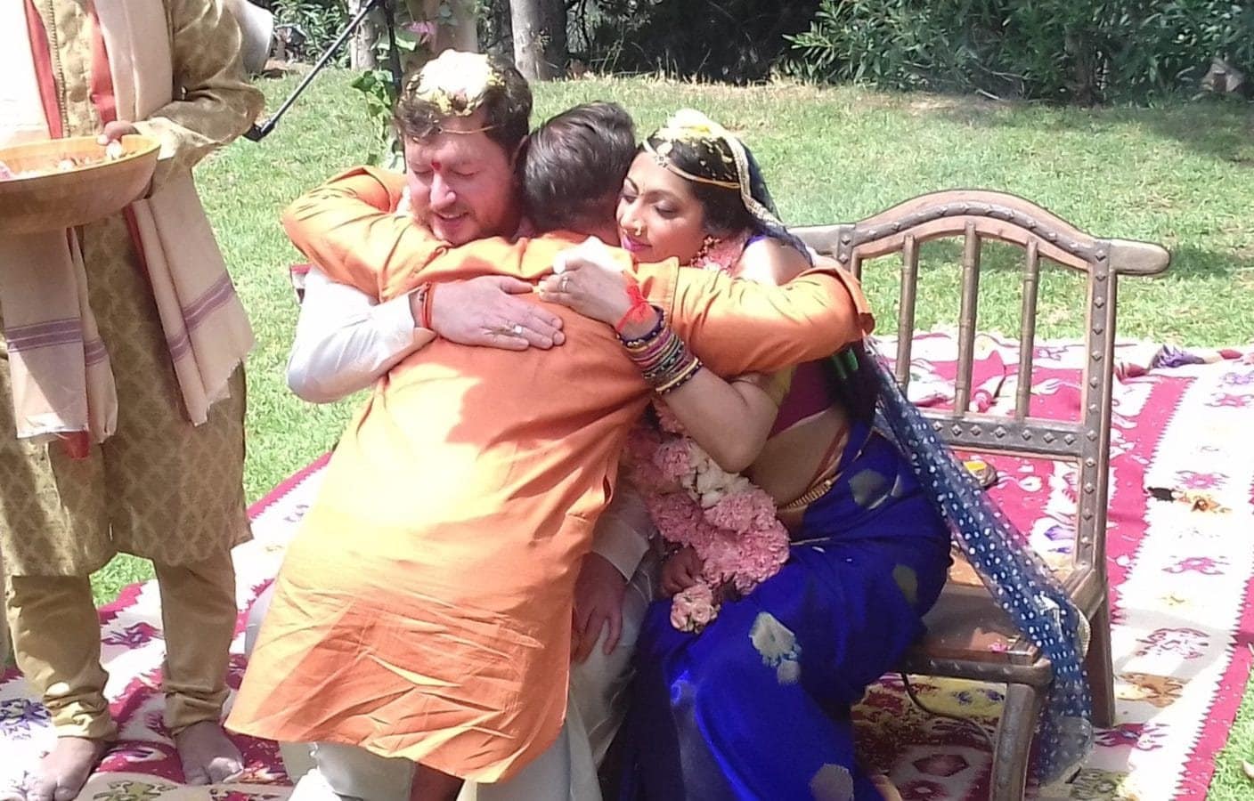 Bride in blue and groom in saffron embracing an elder on the ceremonial mat, with a plate of akṣata held ready beside them