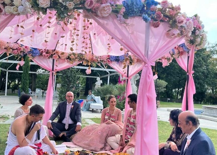 Priest seated before the fire vessel reciting mantras beneath a garlanded pink maṇḍapa, with the couple and both families seated on the lawn