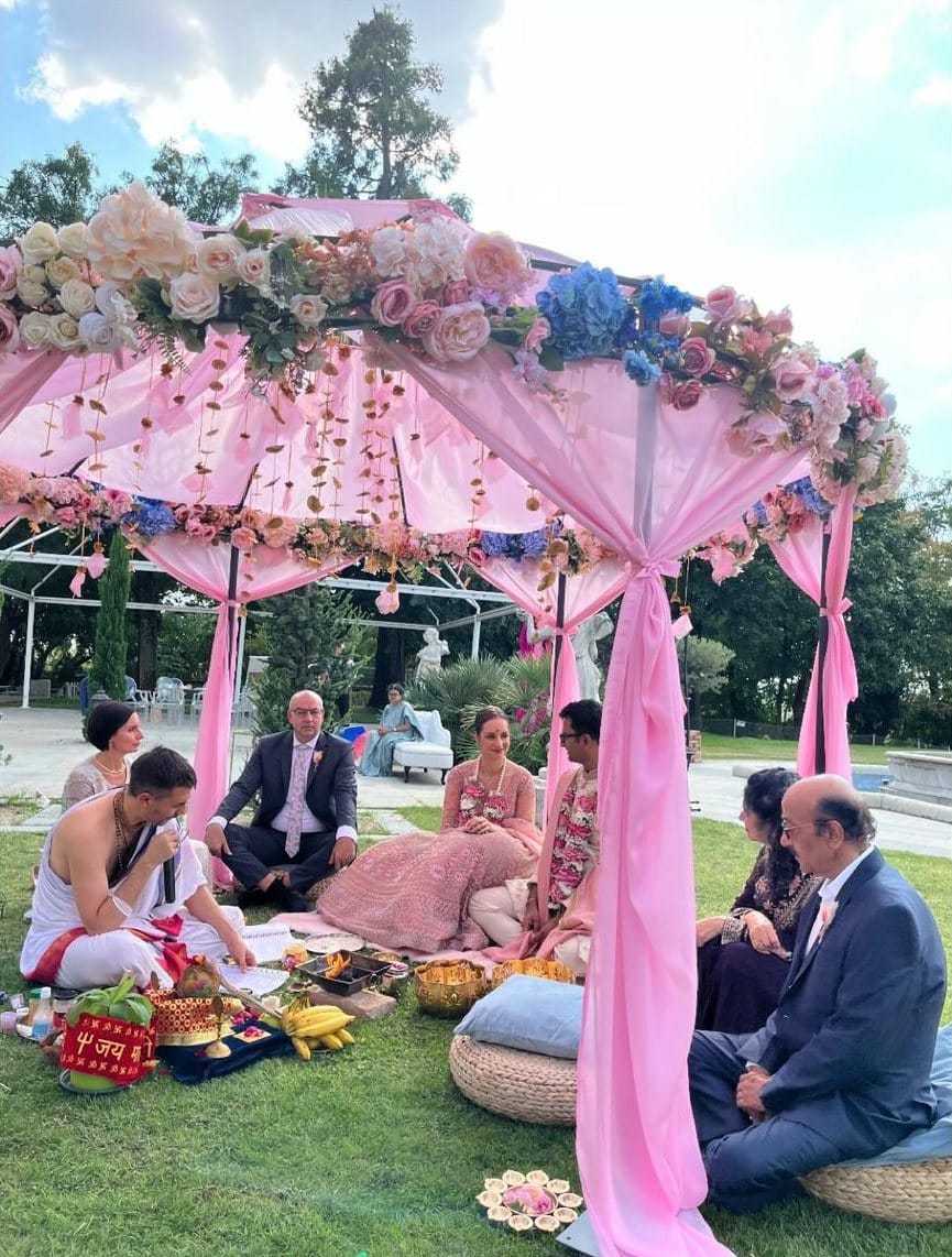 Priest seated before the fire vessel reciting mantras beneath a garlanded mandap, the couple and both families seated on the lawn