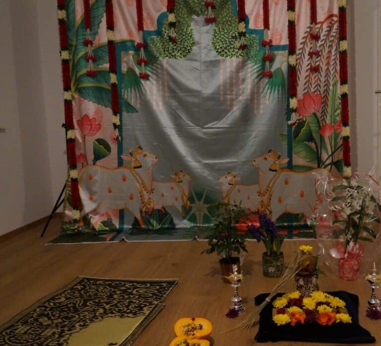 Painted pichwai backdrop of cows and lotuses hung with garlands, with a flower-covered seat, kalaśa and silver lamps
