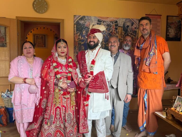 Hindu bride in a red lehenga with mehndi and groom in a white sherwani, both garlanded, with family and Pandit Sahadev