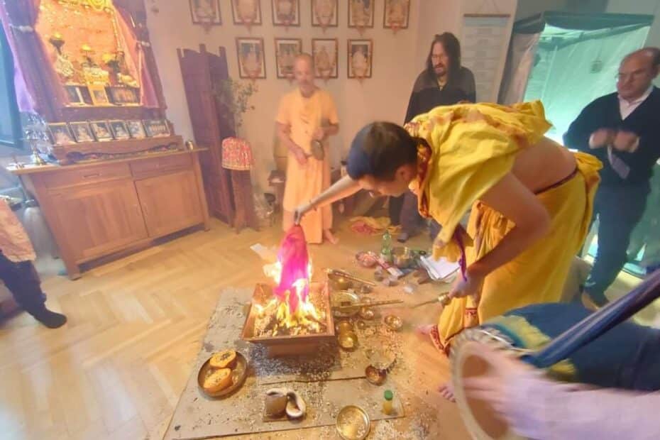 Priest in a yellow dhoti placing a cloth offering into a blazing copper homa kuṇḍa as others stand by