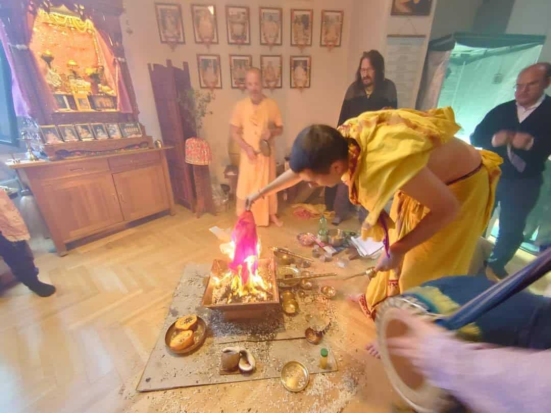 Priest in a yellow dhoti placing a cloth offering into a blazing copper homa kuṇḍa as others stand by