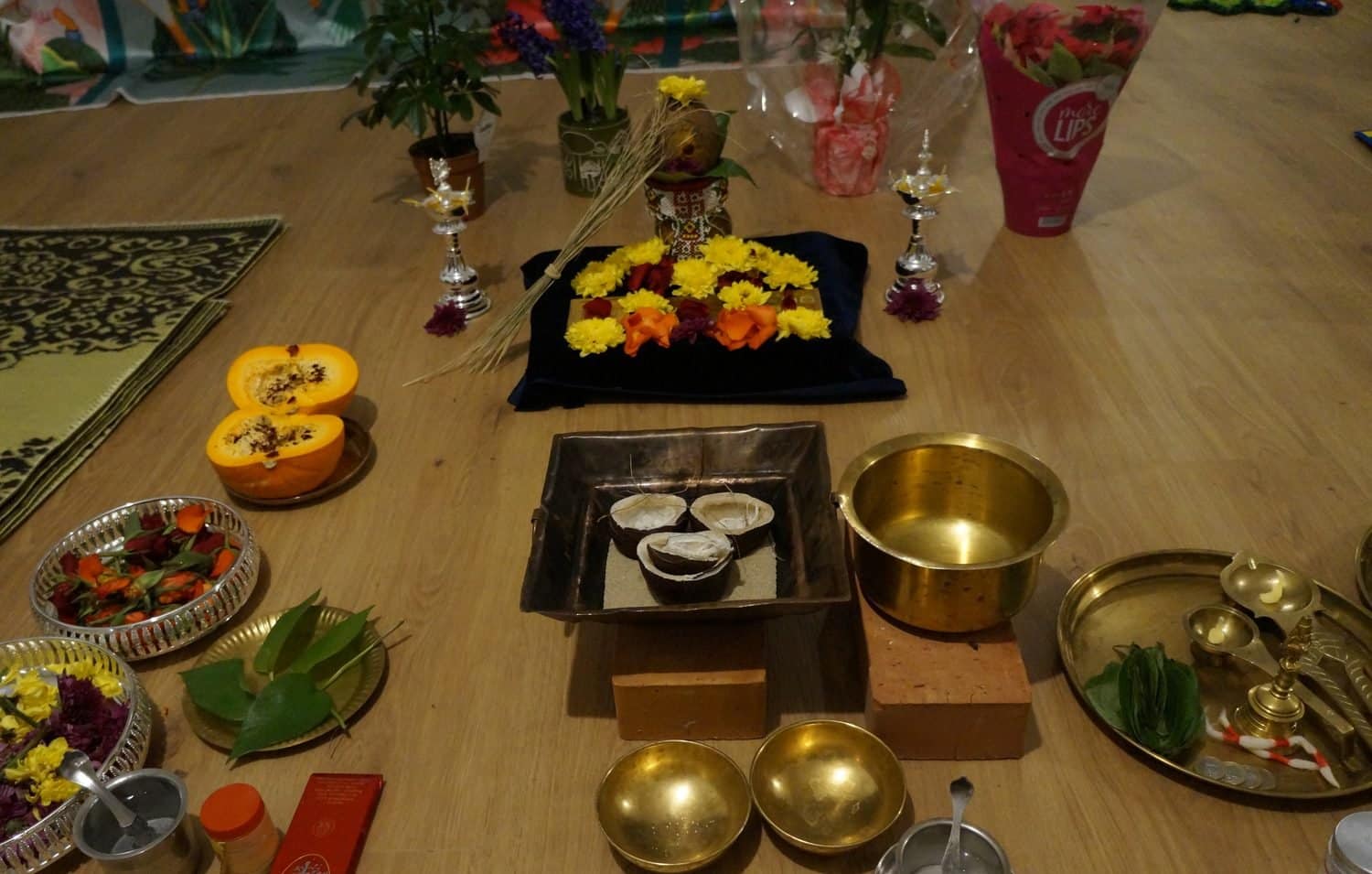 Copper homa kuṇḍa with coconut halves, brass vessels, offering ladles, flowers and silver lamps before a decorated kalaśa