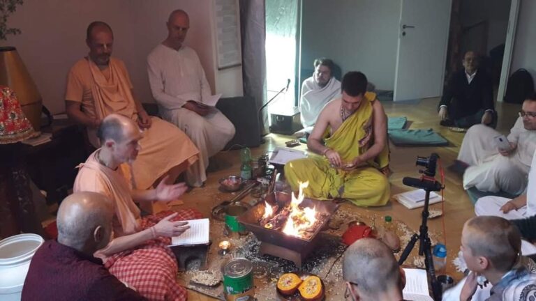 Priest in yellow making offerings at a copper homa kuṇḍa as participants around him follow from printed texts